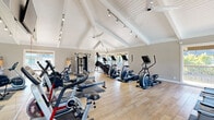 This gym offers a single, generously sized exercise room designed to support a full range of fitness activities. The space is defined by its striking vaulted, cathedral-style ceiling with white-painted shiplap paneling and exposed beams, creating an open, airy atmosphere that feels considerably larger than a typical fitness facility. Track lighting runs along the roofline, supplementing the abundant natural light that pours in through multiple large windows and glass doors, which also provide outdoor views of surrounding greenery. Light wood-tone flooring runs throughout, lending a clean and modern aesthetic. The room is comprehensively equipped with a wide variety of cardio machines, including treadmills, elliptical trainers, stationary bikes, rowing machines, and stair climbers, as well as a multi-function strength training station. Exercise mats and stability balls are also available for stretching and functional training. A large wall-mounted flat-screen television and a wall clock are positioned for easy viewing during workouts. Ceiling fans provide additional air circulation throughout the space. Full-length mirrors along one wall visually expand the room and assist with form during exercise. The overall ambiance is bright, welcoming, and well-organized, making this gym a functional and motivating environment for fitness enthusiasts.