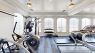 Located at 10 W Minnezona Ave in Phoenix, AZ, this fitness center offers a thoughtfully designed workout space that balances functionality with refined architectural detail. The single-room facility is finished with elegant crown molding, a tray ceiling, and ceiling fans that contribute to a polished, comfortable environment. A row of tall arched windows lines one wall, fitted with frosted glass that allows abundant natural light to fill the space while maintaining privacy. Large mirrors along an adjacent wall visually expand the room and enhance the open, airy feel. The flooring consists of durable, patterned carpet tiles in a cool blue-grey palette, providing both traction and style. Equipment is well-distributed throughout the space, including a treadmill, elliptical trainer, stationary cycle, and a multi-station cable and resistance machine. A tiered dumbbell rack holds a full range of free weights, complemented by stability balls and floor exercise mats for stretching and functional training. A wall-mounted television and clearly marked exit door add practical convenience. Recessed lighting and ceiling-mounted fans ensure the space remains well-lit and ventilated. This fitness center delivers a comprehensive workout environment suited for both cardio and strength training needs.