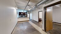 This urban loft-style unit at 144 4th St in Burlington, NC, showcases a compelling industrial aesthetic throughout its six rooms. Polished and stained concrete floors run continuously across the space, complemented by exposed structural beams and bold silver ductwork overhead. The open living and kitchen area features a granite-topped breakfast bar, dark cabinetry, stainless steel appliances, pendant lighting, and a ceiling fan. Floor-to-ceiling windows with horizontal blinds and a glass-paneled exterior door flood the main area with natural light. The bathroom is finished with a granite vanity, dark wood cabinetry, and a tub-shower combination. Additional rooms offer flexible, open layouts with track lighting and clean white walls.