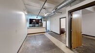 This urban loft-style unit at 144 4th St in Burlington, NC, showcases a compelling industrial aesthetic throughout its six rooms. Polished and stained concrete floors run continuously across the space, complemented by exposed structural beams and bold silver ductwork overhead. The open living and kitchen area features a granite-topped breakfast bar, dark cabinetry, stainless steel appliances, pendant lighting, and a ceiling fan. Floor-to-ceiling windows with horizontal blinds and a glass-paneled exterior door flood the main area with natural light. The bathroom is finished with a granite vanity, dark wood cabinetry, and a tub-shower combination. Additional rooms offer flexible, open layouts with track lighting and clean white walls.