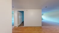 Located at 1 River Place in Lowell, MA, this two-bedroom, one-bathroom unit offers a compact and efficiently designed single-floor layout. The interior features a mix of light hardwood flooring in the main living areas and soft carpeting throughout both bedrooms, creating a comfortable and cohesive aesthetic.

The primary bedroom is a standout space, featuring a distinctive curved bay window framed in dark green that brings in natural light and offers an architectural detail rarely found in similarly sized units. A built-in closet provides practical storage. The second bedroom is equally functional, featuring carpeted flooring, a wall-mounted radiator for heating, and closet access.

The bathroom is clean and well-appointed, featuring a white vanity with chrome fixtures, a mirrored medicine cabinet with overhead lighting, and hardwood flooring that complements the rest of the unit.

The main living area opens into the space with hardwood floors, neutral walls, and windows equipped with roller shades, allowing flexible light control. A wall-mounted HVAC unit and a refrigerator are visible, indicating practical utility features. A connecting hallway ties the rooms together, with crisp white walls and consistent trim throughout. The overall ambiance is clean, neutral, and move-in ready.