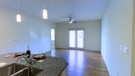 This single-floor residence offers a clean, open layout designed for comfortable everyday living. The main living area is immediately welcoming, bathed in natural light streaming through white-shuttered windows and a set of classic French doors that lead to an outdoor space, creating an airy, connected feel. Smooth, light-toned hardwood floors run throughout, adding warmth and continuity to the space. A ceiling fan is centrally positioned overhead, enhancing both comfort and airflow in the living area. The kitchen flows seamlessly into the living space, featuring elegant granite countertops with a subtle, contrasting pattern. A stylish pendant light hangs above the counter, providing focused task lighting while contributing to the overall modern aesthetic. The walls are finished in a crisp, neutral tone that amplifies the brightness of the space and provides a versatile backdrop for any décor style. The combination of the French doors, abundant window coverage with plantation shutters, and open floor plan gives the space a light-filled, welcoming ambiance that feels both functional and inviting throughout the day.