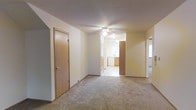 This single-floor residence offers a straightforward and functional layout designed for comfortable everyday living. The main living area is finished with wall-to-wall carpeting in a neutral tone, providing a warm and cohesive feel throughout the space. A ceiling fan with an integrated light fixture is centrally mounted overhead, offering both air circulation and ambient lighting. The room features clean white walls and a textured ceiling, contributing to a simple and understated aesthetic.

Wide doorways connect the living area to adjoining spaces, including what appears to be a kitchen visible in the background, which features wood-toned cabinetry and a window that allows natural light to filter into the interior. A closet door with a warm wood finish is situated along one wall, providing convenient storage. Additional doorways lead to further rooms, suggesting a connected and accessible floor plan.

The open sightlines between rooms create a sense of flow and continuity, making the space feel cohesive despite its modest scale. Natural light from multiple directions brightens the interior, reducing the reliance on artificial lighting. Overall, the space presents a clean, move-in ready environment with practical features suited for a range of occupants.
