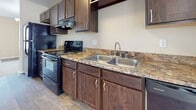 This 1-bedroom, 1-bathroom residence is thoughtfully laid out across a single floor, making efficient use of its space. The kitchen is well-equipped, featuring rich dark wood cabinetry paired with granite-style countertops, a double-basin stainless steel sink, and black appliances including a range and dishwasher. Wood-look plank flooring runs through the kitchen and bathroom, adding warmth and easy maintenance to these functional areas.

The bedroom is generously proportioned with plush wall-to-wall carpeting and warm neutral wall tones. It features a large window with vertical blinds that allow ample natural light, as well as a closet for storage. A second carpeted room, also with neutral finishes, provides additional flexible living space and its own closet access.

The bathroom continues the cohesive design aesthetic with matching dark wood cabinetry, a granite-style vanity countertop, an undermount sink, and wood-look flooring. A tiled shower-tub combination and a grab bar complete the practical layout.

Throughout the unit, consistent warm beige tones, white trim, and clean lines create a cohesive and inviting atmosphere. The layout, as seen in the dollhouse view, is compact yet well-organized, offering comfortable living across all areas.