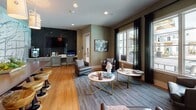 Located at 3 Woodcock Rd in Savannah, GA, this single-floor community space offers a thoughtfully designed interior that blends comfort with contemporary style. The open layout is divided into two distinct yet connected zones, each with its own character and function.

The lounge area is anchored by warm hardwood-style flooring and furnished with a curated mix of leather and upholstered seating, including rounded armchairs and a sofa arranged around paired marble-top coffee tables with gold accents. Large windows dressed in sheer dark drapes flood the space with natural light and offer views of an outdoor pool area. A dark tile-clad feature wall hosts a large flat-screen television alongside a dedicated workspace with cabinetry and a beverage refrigerator, making the space equally suited for relaxation or productivity. Framed artwork and decorative accents add a refined, gallery-like quality to the walls.

The adjacent dining area makes a striking visual statement with a large-scale abstract mural rendered in silver and white tones, depicting what appears to be an artistic city map. Beneath a cluster of cylindrical pendant lights, a dark wood communal table surrounded by industrial-style adjustable stools provides an inviting gathering spot. Together, the two areas create a versatile, design-forward environment ideal for socializing and everyday community use.