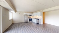This well-appointed single-floor unit offers a clean, modern aesthetic with a thoughtfully designed layout. The open-concept living and kitchen area is anchored by light gray luxury vinyl plank flooring that extends throughout the main living spaces, giving the interior a cohesive and airy feel. Large windows allow natural light to filter in, complementing the neutral cream-toned walls and bright ceiling panels.

The kitchen is a standout feature, outfitted with crisp white shaker-style cabinetry accented by matte black hardware, dark granite-look countertops, and a classic white subway tile backsplash. A full suite of stainless steel appliances — including a refrigerator, electric range, over-the-range microwave, and dishwasher — along with a gold-toned faucet at the sink, adds a contemporary touch.

The living area is generously proportioned, with beamed ceiling details lending subtle architectural character. A front entry door with a sidelight panel provides a welcoming entrance. Bedrooms are finished with plush carpet for comfort and include closet space. The bathroom features a clean white surround shower unit with built-in corner shelving. Throughout the unit, the design balances practicality with a fresh, updated style.