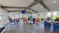 The Eller Oaks Leasing Center is a thoughtfully designed, single-floor office space that combines contemporary style with a welcoming, open atmosphere. Upon entry, visitors are greeted by a bright foyer featuring bold, colorful artwork, sleek flooring in a cool gray wood finish, and a branded Lion Real Estate Group entryway mat that sets a professional tone. The space opens into a spacious main area defined by striking vaulted ceilings with exposed gray-painted beams and a dramatic geometric pendant light fixture that serves as a central design focal point. Recessed lighting complements the abundant natural light streaming in through floor-to-ceiling windows and glass panels that wrap the perimeter, offering unobstructed views of the surrounding greenery and outdoor landscape. The main lounge area features vibrant blue and orange upholstered seating arranged around a central white coffee table, creating an inviting space for residents and prospects. A dedicated workspace with a warm wood-toned desk and coordinating blue seating provides a functional area for leasing consultations. A front reception counter with a dark, modern finish anchors the interior, while additional seating arrangements throughout accommodate varied uses. The overall ambiance is energetic yet professional, with a cohesive color palette, contemporary furnishings, and ample natural light contributing to a polished and welcoming environment.