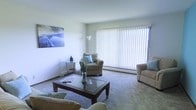 This single-floor residence offers a welcoming and well-lit living environment designed with comfort and practicality in mind. The living room serves as the centerpiece of the space, featuring wall-to-wall carpeting in a neutral tone that adds warmth underfoot. A large sliding glass door fitted with vertical blinds dominates one wall, allowing an abundance of natural light to fill the room while providing access to the outdoors. The light, airy ambiance is complemented by smooth white walls and a clean ceiling, creating a sense of openness throughout. The living area is thoughtfully arranged with seating including a sofa and armchair in complementary beige tones, centered around a dark wood coffee table that grounds the space. Decorative touches such as a floor lamp, a vase with dried botanicals, and wall art depicting a serene landscape add personality without overwhelming the room's understated aesthetic. A small side table with a chair occupies a corner near the window, offering a quiet nook for reading or relaxing. The overall design emphasizes a calm, neutral palette with functional furnishings, making it a comfortable and inviting single-floor living space.
