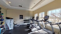 This fitness center is thoughtfully designed to support a comprehensive workout experience. The room features a tray ceiling with recessed lighting and crown molding, lending a polished, finished look uncommon in typical gym spaces. Warm beige walls and dark rubberized flooring create a clean, functional aesthetic throughout.

Natural light is a standout feature, with multiple large windows lining the walls and a set of French doors that open to the outdoors, flooding the space with sunlight and providing views of the surrounding exterior. Full-length mirrors along one wall visually expand the room and assist with form during exercises.

The cardio section is well-stocked with a row of True-brand ellipticals and treadmills, complemented by a rowing machine and a stationary bike. On the strength training side, the space includes a Body-Solid cable machine, a dedicated free weight rack with a full range of dumbbells, a weight bench, and a medicine ball rack stocked with various weighted balls. Resistance bands, exercise mats, and fitness guidelines posted on the wall round out the amenities. The overall layout is organized and spacious, allowing users to move freely between equipment.