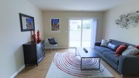 This two-floor residential space offers a well-organized and functional layout designed for comfortable everyday living. The main living area is a standout feature, showcasing warm hardwood-style flooring that extends throughout the room and creates a cohesive, inviting atmosphere. A large sliding glass door serves as the focal point of the living room, flooding the space with natural light and providing a direct view of the surrounding greenery outdoors. The living room is thoughtfully arranged with a contemporary gray sofa, a sleek rectangular coffee table, and a bold patterned area rug that adds a striking visual element to the room. A stylish wire accent chair and a dark-toned dresser with decorative hardware contribute to the eclectic yet coordinated design aesthetic. Colorful wall art enhances the room's personality, complementing the neutral, warm-toned walls that provide a clean and versatile backdrop throughout the space. The overall ambiance is bright, airy, and welcoming, with a modern sensibility balanced by approachable, lived-in charm. Spanning two floors, this space provides ample room for both relaxing and everyday activities, making it a practical and visually appealing place to call home.