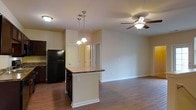 This single-floor residence features an open-concept design that integrates the kitchen and living area into one cohesive space. The kitchen is equipped with dark cabinetry, a black refrigerator, and a peninsula-style island with a stone-look countertop, offering both additional prep space and casual seating potential. A decorative pendant light fixture hangs above the island, providing focused task lighting while adding a stylish touch. The living area extends naturally from the kitchen, finished with warm hardwood-style flooring that runs throughout the space, contributing to a unified and inviting aesthetic. A ceiling fan with a light kit is installed in the living area, ensuring both air circulation and overhead illumination. Neutral beige walls create a clean, versatile backdrop throughout. Doorways visible in the background suggest access to additional rooms, lending functional depth to the layout. The open flow between the kitchen and living space makes the unit well-suited for both everyday living and entertaining. Overall, the space presents a practical and well-appointed interior with consistent finishes throughout.