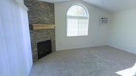 This single-floor home makes a strong impression with its thoughtfully designed living space. The living room serves as the centerpiece, boasting soaring vaulted ceilings that amplify the sense of openness and airiness. A dramatic floor-to-ceiling stacked-stone fireplace with a natural wood mantel anchors the room, adding warmth and a rustic yet refined aesthetic. Plush, neutral-toned carpeting extends across the floor, providing comfort underfoot and contributing to the room's inviting atmosphere. Natural light pours in through a graceful arched window, whose curved silhouette adds an elegant architectural touch while framing views of the surrounding greenery. A wall-mounted air conditioning unit ensures year-round climate comfort. The clean white walls serve as a versatile backdrop, offering a blank canvas that complements a wide range of interior design styles. Together, these elements create a living space that balances casual comfort with distinctive architectural character, making it both functional and visually compelling for everyday living.