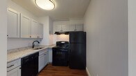 Located at 1 Water St in Haverhill, Massachusetts, this single-floor unit offers a clean, functional layout designed for comfortable everyday living. Rich, warm-toned hardwood-style flooring runs throughout the living areas, creating a cohesive and inviting atmosphere.

The kitchen is efficiently designed, featuring white shaker-style cabinetry that extends to the ceiling for ample storage, granite-patterned countertops, and a full suite of black appliances including an electric range, refrigerator, and dishwasher. An under-cabinet range hood and a deep single-basin sink with a matte black faucet round out the space, all tied together with a textured ceiling and flush-mount lighting overhead.

The bathroom is bright and accessible, equipped with a full soaking tub with a tiled surround, stainless steel grab bars for added safety, a granite-topped vanity, and light tile flooring. Open built-in shelving adjacent to the tub provides convenient storage.

The living areas are open and airy, with crisp white walls, textured ceilings, and large windows that allow natural light to fill the space and offer views of the surrounding streetscape. A wall-unit air conditioner and baseboard heating ensure year-round climate comfort. The overall design is clean and straightforward, making this a practical and move-in-ready space in the heart of Haverhill.