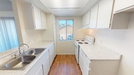 This single-story residence offers a clean, functional layout designed for comfortable everyday living. The galley-style kitchen is a standout feature, lined on both sides with white cabinetry, light speckled countertops, and a double-basin sink. A white electric range sits alongside generous counter space, and a window at the end of the kitchen allows natural light to brighten the room. Warm hardwood-style flooring runs throughout the kitchen, adding contrast to the crisp white finishes.

The living area is open and airy, with plush neutral carpeting underfoot and a textured ceiling overhead. A ceiling fan adds to the comfortable atmosphere, while full-length vertical blinds frame a sliding glass door that invites natural light and provides outdoor access. A pass-through opening connects the living area to the kitchen, enhancing the sense of openness between the two spaces.

The bedroom is quiet and well-lit, featuring the same neutral carpet as the living area and a window dressed with vertical blinds that filter soft, natural light. The bathroom is efficiently laid out with a wide vanity, light countertops, white cabinetry with ample storage below, and a large mirror. The space connects conveniently to the bedroom, completing a cohesive and practical floor plan throughout.