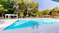 Located at 411 Gregory Dr in Cary, NC, this community recreational facility is centered around a generously sized outdoor swimming pool that serves as the focal point of the space. The pool features clearly marked depth indicators, a stepped entry point, and metal handrails for safe and easy access. The surrounding concrete deck provides ample room for lounge chairs, offering residents plenty of space to relax and sunbathe. A covered gazebo with seating is positioned poolside, providing a shaded retreat for those seeking relief from the sun. The facility is enclosed by a metal perimeter fence, ensuring safety and privacy throughout.

The adjacent single-story clubhouse building is designed with a traditional gabled roofline and light-colored siding accented by brick detailing. Large glass doors and windows along the pool-facing façade allow natural light to flow into the interior while maintaining a visual connection to the outdoor area. A covered entrance portico with white columns adds a welcoming architectural touch to the building's exterior.

Lush, mature trees surround the perimeter of the facility, creating a natural green backdrop that enhances the sense of seclusion. Vibrant flowering shrubs accent the fenced boundary, adding seasonal color to the landscape. A children's playground is visible within the fenced area, making this a well-rounded recreational destination for all ages.