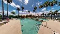 This expansive outdoor pool area offers a resort-style retreat surrounded by towering palm trees that create a lush, tropical ambiance beneath open blue skies. The centerpiece is a generously sized swimming pool with clear, turquoise water, complemented by two raised spa or water feature platforms integrated seamlessly into the pool design. The wide concrete deck surrounding the pool provides ample room for lounge seating and relaxation, with multiple deck chairs positioned throughout the area. A covered patio structure with arched architectural detailing and a terracotta tile roof anchors one side of the pool area, offering shaded respite. An additional utility or amenity building is situated nearby, providing convenient access to pool facilities. The entire area is enclosed by a sturdy wrought-iron fence with gated entry points, ensuring safety and security. Classic lamp posts are positioned along the walkways, adding a charming, well-maintained character to the space. Views of the surrounding greenery and a glimpse of a playground area beyond the fence add to the community-oriented atmosphere. The overall setting combines functional outdoor amenities with a vibrant, sun-filled environment ideal for leisure and recreation.