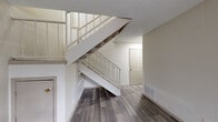 This two-floor residence offers a well-organized layout with distinct living spaces across both levels. The main floor is anchored by a spacious living area finished with durable wood-look flooring that extends throughout. A standout feature is the floor-to-ceiling stacked stone fireplace with a wood mantel, which serves as a dramatic focal point. Large windows and a sliding glass door flood the living area with natural light while providing views to the outdoors. An open-riser staircase with white metal railings connects the main floor to the upper level, adding an architectural element that enhances the sense of openness. The kitchen is efficiently designed, featuring gray cabinetry, granite-style countertops, and black appliances including a full-size refrigerator and range. A bathroom on the main level includes a dark-toned vanity with ample cabinet storage and a large mirror. The upper level transitions to plush carpeting and includes an open loft-style area with a ceiling fan and railed overlook. A textured ceiling is consistent throughout the home, contributing to its classic residential character. The overall palette of neutral tones and clean finishes creates a bright, functional, and comfortable living environment.