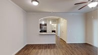 This single-floor residence offers a thoughtfully designed open-concept layout that balances style and functionality. Rich hardwood-style flooring flows consistently throughout the main living areas, lending warmth and visual continuity to the space.

The living area is bright and airy, highlighted by elegant crown molding along the ceiling and a classic ceiling fan with a light fixture that ensures year-round comfort. Vertical blinds on the sliding glass door allow for flexible light control and access to the outdoors.

A graceful arched passageway serves as the defining architectural feature of the space, connecting the living area to the kitchen in a refined and open manner. The kitchen is well-equipped, showcasing dark cabinetry that contrasts beautifully against light granite-style countertops. A breakfast bar peninsula provides casual seating and additional workspace, while pendant lighting above adds a stylish, modern touch. Stainless steel appliances, including a refrigerator, complete the kitchen's polished look.

Beyond the kitchen, a hallway leads to additional rooms, including what appears to be a bathroom with visible vanity cabinetry. Paneled interior doors with arched detailing reinforce the cohesive architectural character found throughout. Overall, the space presents a clean, move-in-ready aesthetic with tasteful finishes throughout.