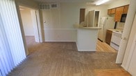 This single-floor residence offers a straightforward and functional layout centered around an open-concept living and kitchen area. The living space features wall-to-wall carpeting in a neutral beige tone, providing a warm underfoot feel, while the walls are finished in a clean, light color palette that keeps the space feeling bright and airy. A chair-rail-style ledge runs along the lower portion of the main wall, adding a subtle architectural detail to the room. The kitchen is defined by a peninsula counter with a laminate top that extends into the living area, creating a natural divide between the two spaces while maintaining an open sightline. The kitchen transitions to smooth hardwood-style flooring, offering a practical surface for food preparation and daily use. A wood-toned upper cabinet and a full-size white refrigerator are visible, contributing to the kitchen's classic and utilitarian character. A closet with a louvered door is accessible from the living area, offering convenient built-in storage. Overhead fluorescent lighting keeps the interior well-illuminated throughout. The overall ambiance is clean, neutral, and move-in ready, with a practical design suited for comfortable day-to-day living.