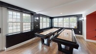 Located at 201 S Huntington Ave in Boston's Jamaica Plain neighborhood, this single-floor amenity space blends bold design with functional recreational features. The interior is defined by striking dark textured wallcoverings that contrast sharply with crisp white trim and ceilings, creating a sophisticated, moody aesthetic throughout.

The lounge area offers a welcoming setting with warm hardwood flooring, a caramel-toned sofa, a modern round coffee table, and contemporary accent chairs arranged on a cowhide-style rug. Large multi-pane windows wrap the room, flooding the space with natural light and providing views of the surrounding neighborhood. A glass door leads directly to an outdoor area, enhancing connectivity between indoor and outdoor spaces.

A central corridor with clean white walls and recessed lighting connects the lounge to the game room, where the color palette shifts with bold accent walls in red and navy, adding a playful energy. The game room is a standout feature, housing two full-size shuffleboard tables positioned beneath expansive windows that overlook an outdoor terrace. The dark wall treatment continues here, reinforcing the cohesive design language of the space.

Throughout, hardwood floors, recessed lighting, and large windows unify the areas into a well-appointed, light-filled space ideal for social gatherings and recreation.