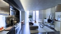 This contemporary single-floor space showcases a thoughtfully designed open-plan layout that seamlessly blends the kitchen, dining, and living areas into one cohesive environment. The exposed concrete ceiling adds an industrial-chic character, complementing the dark hardwood flooring and monochromatic color palette throughout.

The kitchen is sleek and modern, fitted with dark cabinetry, integrated appliances, and a compact dining counter with seating — ideal for casual meals. It flows naturally into the living area, which is anchored by a minimalist sofa, a glass coffee table, and a plush area rug, creating a comfortable yet refined atmosphere.

Floor-to-ceiling windows span the far wall, flooding the space with abundant natural light and offering expansive urban views of the surrounding cityscape. A large indoor plant adds a welcome touch of greenery, softening the clean, architectural lines of the interior. A lounge chair positioned near the windows creates an inviting reading or relaxation nook.

The overall ambiance is modern, calm, and urban — a well-curated space that balances functional living with sophisticated design. The use of neutral tones, quality materials, and carefully chosen furnishings gives the space a polished, gallery-like feel.