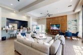Located at 11000 Point Sylvan Circle in Orlando, FL, this single-floor community clubhouse offers a thoughtfully designed gathering space that blends modern aesthetics with functional amenities. The main lounge serves as the heart of the space, showcasing tray ceilings with ceiling fans, large multi-pane windows framed by sheer white curtains, and a striking navy blue accent wall that adds depth and character. Wood-look tile flooring runs throughout, complementing a bold wood-paneled feature wall that houses a large flat-screen television and media shelving. Plush neutral sofas and armchairs are arranged around coffee tables, creating a welcoming environment for socializing. A reception desk area with upholstered guest seating provides a professional point of contact. Adjacent to the lounge, a casual bar-height counter with mosaic tile backsplash adds a social gathering spot. A secondary room, accessible through glass-paneled French doors, features a round dining table with mid-century modern blue chairs, a built-in wood bookcase, and workstation desks — ideal as a business center or private meeting space. A contemporary multi-bulb chandelier adds a stylish focal point overhead. Natural light floods the entire space, creating a bright, open, and inviting ambiance throughout.
