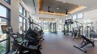 This single-level fitness center is designed to support a comprehensive workout experience within a bright, modern space. The layout is generously sized, accommodating a wide variety of equipment zones without feeling crowded. Architecturally, the facility features soaring ceilings with elegant tray detailing, warm recessed lighting accented by ambient cove illumination, and multiple ceiling fans that contribute to a comfortable environment during exercise.

Floor-to-ceiling windows and oversized black-framed glass panels line the perimeter, flooding the interior with abundant natural light while offering views of a resort-style pool, manicured lawns, and tropical palm trees. The flooring throughout is durable, cushioned gym carpeting suited for both cardio and strength activities.

The cardio section is well-appointed with treadmills, StairMaster steppers, ellipticals, and Les Mills cycling bikes. A separate area houses a functional training rig with weight plates and barbells, cable machines, rowing machines, and a plate-loaded weight rack. Exercise balls, dumbbells, and benches are neatly organized along a textured accent wall. Large wall-mounted televisions provide entertainment throughout. The overall ambiance is modern, airy, and resort-inspired.