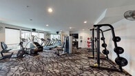 This fitness center in College Station, Texas, offers a comprehensive workout environment spread across a single floor. The space is thoughtfully designed with a patterned, textured carpet that adds visual interest while providing a comfortable surface underfoot. Recessed lighting throughout the ceiling ensures bright, even illumination, complemented by large windows along one wall that invite natural light and provide outdoor views.

The cardio zone is a standout feature, housing a row of modern treadmills, stair climbers, and additional aerobic machines arranged in an orderly fashion. A large wall-mounted mirror runs alongside this area, visually expanding the space and allowing users to monitor their form. Wall-mounted fans are positioned strategically for added comfort, and a flat-screen television is mounted for entertainment during workouts.

The strength training area includes a power rack with a barbell and weight plates, a decline bench, and a cable machine, offering versatile options for resistance training. A dedicated section houses a tiered dumbbell rack stocked with a full range of free weights, positioned near a windowed door that provides additional natural light. A pass-through window connects to an adjacent area where medicine balls and additional equipment are visible. The overall ambiance is clean, modern, and functional.
