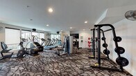 This fitness center in College Station, Texas, offers a comprehensive workout environment spread across a single floor. The space is thoughtfully designed with a patterned, textured carpet that adds visual interest while providing a comfortable surface underfoot. Recessed lighting throughout the ceiling ensures bright, even illumination, complemented by large windows along one wall that invite natural light and provide outdoor views.

The cardio zone is a standout feature, housing a row of modern treadmills, stair climbers, and additional aerobic machines arranged in an orderly fashion. A large wall-mounted mirror runs alongside this area, visually expanding the space and allowing users to monitor their form. Wall-mounted fans are positioned strategically for added comfort, and a flat-screen television is mounted for entertainment during workouts.

The strength training area includes a power rack with a barbell and weight plates, a decline bench, and a cable machine, offering versatile options for resistance training. A dedicated section houses a tiered dumbbell rack stocked with a full range of free weights, positioned near a windowed door that provides additional natural light. A pass-through window connects to an adjacent area where medicine balls and additional equipment are visible. The overall ambiance is clean, modern, and functional.