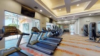 Located at 2450 Lake Debra Dr in Orlando, Florida, this fitness center offers a well-equipped, single-floor space designed to support a full range of workout routines. The facility features a spacious main exercise area with an impressive lineup of cardio machines, including multiple treadmills and elliptical trainers by Matrix, as well as stationary bikes. A dedicated strength training zone houses a variety of resistance machines, cable systems, TRX suspension trainers, a punching bag, and a dumbbell rack, providing options for both machine-based and functional training. The flooring transitions between hardwood-style surfaces and patterned carpet with bold orange accents, adding visual energy to the space. Architectural details include a coffered ceiling with recessed lighting, which creates a bright and open atmosphere. Large windows along the perimeter allow natural light to fill the room and offer views of the outdoor surroundings. Wall-mounted flat-screen TVs are positioned for visibility from the cardio equipment. A motivational fitness-themed mural decorates one wall, reinforcing the active atmosphere. A water fountain station is conveniently located near the entrance, and glass doors provide a view into an adjacent common area. The overall design is modern, functional, and energizing.