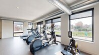 Located at 540 Broadway in Saint Paul, Minnesota, this well-appointed fitness center offers a generously sized, single-floor exercise space designed to support a full range of workout routines. The layout follows an L-shaped floor plan, providing distinct zones for cardio training and strength work, allowing users to move efficiently between equipment types.

The cardio area is lined with floor-to-ceiling windows that flood the space with natural light and frame sweeping views of the Saint Paul cityscape — an energizing backdrop for treadmills, elliptical trainers, and stationary bikes arranged in a neat row. Exposed silver ventilation ductwork runs along the ceiling, lending the space an industrial-modern aesthetic that complements the clean white walls and durable speckled flooring throughout.

The strength training section is equally well-equipped, featuring multiple cable and resistance machines, a multi-function cable tower, adjustable weight benches, and a full dumbbell rack. A built-in hydration station is conveniently mounted on the wall, supporting longer workout sessions. Recessed lighting provides consistent illumination across both zones.

The overall ambiance is modern, bright, and functional — a professional-grade facility that balances practicality with an inviting, open atmosphere suited for daily fitness use.
