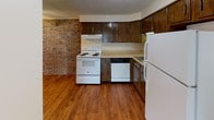 This single-floor residential unit offers a practical and comfortable layout across several well-defined spaces. The kitchen is a standout area, characterized by a striking exposed brick accent wall that adds warmth and texture to the room. Dark wood cabinetry lines the walls, providing ample storage, while wood-look flooring ties the space together. White appliances — including a coil-top electric range, dishwasher, and refrigerator — are neatly arranged in an efficient L-shaped configuration with a sink and generous counter space.

The living area is open and spacious, featuring neutral-toned carpet and a textured ceiling. A pass-through architectural detail connects the living space to adjacent areas, adding visual interest and an open feel. The bedroom features the same soft carpeting, a large window dressed with vertical blinds that allows for natural light and outdoor views, and a closet with bi-fold doors for convenient storage.

The bathroom is bright and clean, finished with light tile flooring and a white vanity with brushed nickel hardware. A full tub and shower enclosure equipped with grab bars provides accessibility, and a mirrored medicine cabinet completes the space. Throughout the unit, a consistent neutral color palette and textured ceilings create a cohesive, understated ambiance.