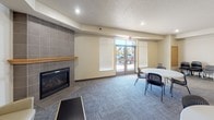 This community room is a well-appointed, single-floor shared space designed to accommodate a variety of group activities and social gatherings. The main area is generously sized, featuring patterned carpet flooring and recessed lighting throughout. A tile-surround fireplace with a wood mantel serves as a focal point, adding warmth to the lounge area, which includes a red leather loveseat and a neutral upholstered armchair. Multiple round tables with stackable chairs are arranged across the open floor, offering flexible seating configurations suitable for meetings, dining, or events. Large windows fitted with roller shades and blinds allow natural light to filter in, while a set of glass double doors provides direct access to the exterior, offering views of the surrounding outdoor area. A fully equipped kitchen is integrated into the space, featuring light wood cabinetry extending to the ceiling, a coil-burner electric range, dishwasher, double sink, and ample counter space — making it well-suited for catered events or communal meal preparation. A second set of wood double doors connects to additional interior areas. The overall ambiance is functional yet comfortable, balancing practicality with a welcoming atmosphere.