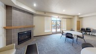 This community room is a well-appointed, single-floor shared space designed to accommodate a variety of group activities and social gatherings. The main area is generously sized, featuring patterned carpet flooring and recessed lighting throughout. A tile-surround fireplace with a wood mantel serves as a focal point, adding warmth to the lounge area, which includes a red leather loveseat and a neutral upholstered armchair. Multiple round tables with stackable chairs are arranged across the open floor, offering flexible seating configurations suitable for meetings, dining, or events. Large windows fitted with roller shades and blinds allow natural light to filter in, while a set of glass double doors provides direct access to the exterior, offering views of the surrounding outdoor area. A fully equipped kitchen is integrated into the space, featuring light wood cabinetry extending to the ceiling, a coil-burner electric range, dishwasher, double sink, and ample counter space — making it well-suited for catered events or communal meal preparation. A second set of wood double doors connects to additional interior areas. The overall ambiance is functional yet comfortable, balancing practicality with a welcoming atmosphere.