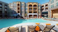 Located at 11701 Mira Lago Blvd in Farmers Branch, Texas, this well-appointed single-floor residence is part of a striking Mediterranean-inspired community. The building's exterior features warm earth tones, arched architectural details, and private balconies set against a beautifully maintained resort-style pool deck complete with lounge chairs and shaded seating areas — perfect for outdoor relaxation under the Texas sun.

Inside, the unit showcases an open-concept layout with wood-style plank flooring that flows seamlessly throughout the living, dining, and kitchen areas. The kitchen is a standout feature, equipped with dark shaker-style cabinetry, stainless steel appliances including a refrigerator, range, and microwave, and a generous granite-topped island that doubles as a breakfast bar with stylish pendant lighting overhead.

The dining area connects fluidly to a comfortable living space, which benefits from natural light streaming through large windows dressed with sheer curtains. A cozy nook with a bistro-style table provides a versatile flex area near the bedroom. The bedroom is accessible directly from the living area and enjoys its own window views. The entry foyer offers convenient closet storage and connects to the kitchen for a practical, well-organized flow throughout the home.