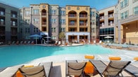 Located at 11701 Mira Lago Blvd in Farmers Branch, Texas, this well-appointed single-floor residence is part of a striking Mediterranean-inspired community. The building's exterior features warm earth tones, arched architectural details, and private balconies set against a beautifully maintained resort-style pool deck complete with lounge chairs and shaded seating areas — perfect for outdoor relaxation under the Texas sun.

Inside, the unit showcases an open-concept layout with wood-style plank flooring that flows seamlessly throughout the living, dining, and kitchen areas. The kitchen is a standout feature, equipped with dark shaker-style cabinetry, stainless steel appliances including a refrigerator, range, and microwave, and a generous granite-topped island that doubles as a breakfast bar with stylish pendant lighting overhead.

The dining area connects fluidly to a comfortable living space, which benefits from natural light streaming through large windows dressed with sheer curtains. A cozy nook with a bistro-style table provides a versatile flex area near the bedroom. The bedroom is accessible directly from the living area and enjoys its own window views. The entry foyer offers convenient closet storage and connects to the kitchen for a practical, well-organized flow throughout the home.