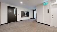 This hallway at The Overlook Residences, located at 7 Erskine Lane in Victoria, BC, serves as a well-appointed residential corridor that combines functionality with thoughtful design. The space is finished with neutral-toned, textured carpeting throughout, complemented by crisp white walls and dark baseboards that create a clean, contemporary aesthetic.

A standout feature of the hallway is a striking branded accent wall clad in vertical wood slat paneling, prominently displaying The Overlook Residences signage in teal and gold — adding warmth and a sense of identity to the corridor. Recessed ceiling lighting provides consistent, even illumination throughout the space, while security cameras and a smoke detector reflect a commitment to resident safety.

Resident suite doors, such as Unit 202, are clearly marked with modern unit number plaques, and elevator access is conveniently located within the corridor. A large framed photographic artwork of a forest road adorns one wall, adding a decorative touch that elevates the ambiance beyond a typical hallway. A window at one end of the corridor introduces natural light, enhancing the overall brightness of the space. The layout is efficient and well-proportioned, offering smooth circulation between units and building amenities.