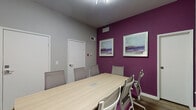 Located at 1700 N DuPont Highway in Dover, Delaware, this single-room conference space is thoughtfully designed to support professional meetings and collaborative work. The room is anchored by a large, light wood rectangular conference table surrounded by ergonomic mesh-back chairs with purple upholstered seats, providing comfortable seating for up to eight participants. A striking deep purple accent wall serves as the room's visual focal point, adorned with framed abstract landscape artwork that adds a creative and polished touch to the environment. The remaining walls are finished in a clean, neutral light gray, creating a balanced and professional atmosphere. Multiple windows along the perimeter allow natural light to filter into the space, complemented by a modern overhead linear lighting fixture that ensures consistent illumination throughout. Wood-look flooring runs across the room, adding warmth and texture underfoot. A built-in cable management port is integrated into the table surface, facilitating seamless connectivity for presentations and meetings. Additional artwork, including a vibrant cityscape panoramic print, decorates the gray walls and adds visual interest. The room is equipped with HVAC vents for climate control and features two interior access doors, offering functional flexibility for the space.