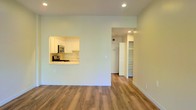This single-floor space offers a clean, open layout with a cohesive design that maximizes functionality. The main living area is finished with warm-toned hardwood-style flooring that runs continuously throughout, lending a sense of unity and spaciousness to the interior. Neutral, light green-toned walls and recessed ceiling lighting create a bright, welcoming ambiance.

A pass-through window connects the living area to the kitchen, which features white cabinetry, a gas range, a built-in microwave, and a sink positioned beneath the pass-through — an efficient layout that keeps the space feeling open while maintaining a defined cooking zone. The kitchen is compact yet well-organized, making practical use of its footprint.

A hallway visible from the main living area leads to additional spaces, including a built-in shelving unit that offers generous open storage. A white refrigerator is also visible, integrated neatly into the layout. The overall design is minimal and functional, with clean lines and a neutral palette that provides a versatile backdrop suitable for a variety of furnishing styles.