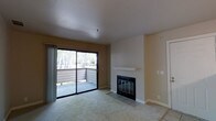 This single-floor space centers around a bright and functional living area designed with comfort and practicality in mind. The room is finished with neutral beige walls and carpeted flooring, creating a warm and understated aesthetic throughout.

A standout feature of the living area is the fireplace, framed by a clean white mantel and set against a tiled surround. It serves as a natural focal point for the room and adds a cozy, inviting element to the overall ambiance.

Natural light flows generously into the space through a large sliding glass door, which opens directly onto a private balcony. The balcony is enclosed by a wooden railing and offers pleasant views of surrounding trees, bringing a sense of the outdoors into the interior. The sliding door is dressed with vertical window treatments that allow for privacy control while maintaining access to the natural light.

The layout is open and unencumbered, offering flexibility for a variety of furniture arrangements. White baseboards and a smooth ceiling contribute to a clean, well-maintained finish throughout. Overall, the space presents a comfortable and livable environment with functional features and a connection to the outdoors.
