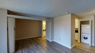 This single-floor space presents a clean, functional layout highlighted by dark hardwood-style flooring that runs throughout, offering a warm contrast to the light, neutral-toned walls. The main living area is open and spacious, with two-tone wall coloring — a warm tan accent wall paired with crisp white on the adjacent side — creating a subtle sense of visual depth. White baseboards provide a clean finish along the floor perimeter. The textured popcorn ceiling adds a classic touch overhead, while a smoke detector is neatly mounted toward the corner. A structural beam or partial wall divides the space slightly, adding architectural character without fully closing off the area. Toward the back, an open doorway leads into what appears to be a bathroom or vanity area, warmly illuminated by a chandelier-style light fixture above a white-cabinetry vanity and dark countertop, offering an inviting contrast to the cooler tones of the main room. Electrical outlets are positioned thoughtfully along the walls. Overall, the space conveys a tidy, move-in ready atmosphere with a neutral palette suitable for a variety of interior styles.