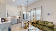 This thoughtfully designed single-floor unit showcases a seamless open-concept layout connecting the kitchen and living area, bathed in natural light from expansive floor-to-ceiling windows. The soaring ceilings amplify the sense of openness throughout, complemented by polished concrete flooring and a crisp white color palette.

The kitchen is a standout feature, equipped with stainless steel appliances including a refrigerator, range with microwave, dishwasher, and a wine cooler. White cabinetry with gold hardware pairs elegantly with granite countertops and a subway tile backsplash. A central island with bar seating provides additional prep space and casual dining, illuminated by stylish globe pendant lights.

The living area flows naturally from the kitchen, offering generous space and direct access to an outdoor view through large windows dressed with sheer curtains and blinds. A ceiling fan keeps the space comfortable.

A bright, art-lined hallway connects the living space to the private bedroom, which features a tray ceiling with recessed lighting and a warm, serene ambiance. Built-in storage solutions are integrated along the hallway corridor.

The bathroom is well-appointed with a granite-topped vanity, gold hardware accents, a large framed mirror, and a marble-tiled shower and tub combination, delivering a clean and polished finish throughout.