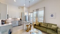 This thoughtfully designed single-floor unit showcases a seamless open-concept layout connecting the kitchen and living area, bathed in natural light from expansive floor-to-ceiling windows. The soaring ceilings amplify the sense of openness throughout, complemented by polished concrete flooring and a crisp white color palette.

The kitchen is a standout feature, equipped with stainless steel appliances including a refrigerator, range with microwave, dishwasher, and a wine cooler. White cabinetry with gold hardware pairs elegantly with granite countertops and a subway tile backsplash. A central island with bar seating provides additional prep space and casual dining, illuminated by stylish globe pendant lights.

The living area flows naturally from the kitchen, offering generous space and direct access to an outdoor view through large windows dressed with sheer curtains and blinds. A ceiling fan keeps the space comfortable.

A bright, art-lined hallway connects the living space to the private bedroom, which features a tray ceiling with recessed lighting and a warm, serene ambiance. Built-in storage solutions are integrated along the hallway corridor.

The bathroom is well-appointed with a granite-topped vanity, gold hardware accents, a large framed mirror, and a marble-tiled shower and tub combination, delivering a clean and polished finish throughout.