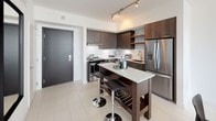 Located in the heart of Miami at 86 SW 8 Street, this modern single-floor residence offers a thoughtfully designed open layout with a clean, contemporary aesthetic throughout. Large-format white porcelain tile floors run seamlessly across the living spaces, complementing the crisp white walls and creating a bright, cohesive atmosphere.

The living area is anchored by expansive floor-to-ceiling sliding glass doors that open onto a private balcony, flooding the interior with natural light and offering sweeping views of the Miami skyline. A ceiling fan and wall-mounted television enhance the functionality of the space.

The kitchen is outfitted with rich dark wood cabinetry, quartz countertops, stainless steel appliances including a French door refrigerator and dishwasher, and a central island with bar seating — ideal for both everyday use and entertaining. Open shelving adds a modern touch.

The bedroom continues the floor-to-ceiling glass motif, with direct balcony access and generous natural light. A dedicated bathroom features marble-look tile surrounds, a combined tub and shower, and modern wood-toned vanity cabinetry. A dedicated laundry area with a stacked full-size washer and dryer adds everyday convenience to this well-appointed Miami residence.