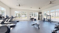 This well-appointed fitness facility is organized across two dedicated exercise rooms, offering a comprehensive environment for a wide range of workout routines. The main exercise room is expansive and airy, featuring high ceilings, recessed lighting, and dual ceiling fans that promote comfortable airflow throughout. Large multi-pane windows line the walls, flooding the space with natural light and providing pleasant views of the surrounding outdoor area, including a pool. The durable rubber flooring provides excellent support underfoot, and a wall-mounted flat-screen TV adds a multimedia element to workouts. Equipment includes treadmills, ellipticals, cable machines, a rowing machine, a weight bench, and a rack of dumbbells, catering to both cardio and strength training needs. A full-length mirror spans one wall, enhancing the sense of space and supporting form-focused training. The second room offers a more intimate setting, equipped with spin bikes, stability balls, and additional dumbbells, along with its own wall-mounted TV — ideal for cycling sessions or focused training. Glass doors provide direct access to the outdoor pool area, seamlessly connecting indoor fitness with the outdoor amenities. Together, the two rooms create a versatile, light-filled facility suited to a variety of fitness goals.