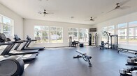 This well-appointed fitness facility is organized across two dedicated exercise rooms, offering a comprehensive environment for a wide range of workout routines. The main exercise room is expansive and airy, featuring high ceilings, recessed lighting, and dual ceiling fans that promote comfortable airflow throughout. Large multi-pane windows line the walls, flooding the space with natural light and providing pleasant views of the surrounding outdoor area, including a pool. The durable rubber flooring provides excellent support underfoot, and a wall-mounted flat-screen TV adds a multimedia element to workouts. Equipment includes treadmills, ellipticals, cable machines, a rowing machine, a weight bench, and a rack of dumbbells, catering to both cardio and strength training needs. A full-length mirror spans one wall, enhancing the sense of space and supporting form-focused training. The second room offers a more intimate setting, equipped with spin bikes, stability balls, and additional dumbbells, along with its own wall-mounted TV — ideal for cycling sessions or focused training. Glass doors provide direct access to the outdoor pool area, seamlessly connecting indoor fitness with the outdoor amenities. Together, the two rooms create a versatile, light-filled facility suited to a variety of fitness goals.