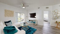This well-designed single-floor residence offers a cohesive layout connecting key living spaces through a clean, contemporary aesthetic. The living room is an immediate standout, featuring soaring vaulted ceilings fitted with a ceiling fan, and wood-look flooring that extends throughout the unit. Natural light pours in through a full-width sliding glass door that opens to a private patio overlooking lush greenery, creating a seamless indoor-outdoor connection. A wall-mounted television and mirrored media console anchor the main wall, while the open layout promotes an airy, spacious feel.

Adjacent to the living area, a dedicated dining space connects to a well-appointed kitchen outfitted with white cabinetry, light countertops, and a full suite of appliances including a refrigerator, electric range, built-in microwave, and dishwasher. Track lighting above provides focused illumination throughout.

The bedroom is a calm, light-filled retreat with a large window framing outdoor greenery, neutral tones, and consistent wood-look flooring. A hallway leads to the full bathroom, which includes a single-sink vanity with a large mirror, a soaking tub with tile surround, and louvered closet doors offering practical storage.
