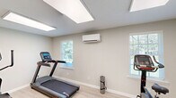 Located at 710 Lynn Dr SE in Jacksonville, Alabama, this dedicated exercise room offers a well-appointed fitness space designed for effective cardio workouts. The room features light wood-look flooring that lends a clean, modern aesthetic throughout. Soft sage-green walls create a calm and focused atmosphere, complemented by bright flush-mounted LED panel lighting in the ceiling that ensures excellent visibility at any time of day. Natural light filters in through multiple windows fitted with white plantation-style blinds, adding an airy quality to the space. A wall-mounted mini-split HVAC unit provides individual climate control, keeping the room comfortable during intense workouts. The fitness center is equipped with several cardio machines, including a treadmill, an elliptical trainer, a stationary upright bike, and an additional multi-function cardio unit, offering a variety of workout options. Two white paneled doors provide entry and access points, and clean white baseboards frame the perimeter of the room. A posted fitness center rules placard is visible on the wall, indicating this space is part of a shared or community fitness facility. The overall design is functional, tidy, and purpose-built for physical activity.
