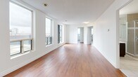 Located at 677 Richmond St in downtown London, Ontario, this well-appointed 2-bedroom, 1-bathroom unit offers a thoughtfully designed single-floor layout. Warm hardwood-style flooring flows throughout the living spaces, complemented by crisp white walls that create a clean, modern aesthetic. The spacious living room is bathed in natural light through large windows and opens toward the unit's entry corridor, providing a welcoming sense of openness. The primary bedroom is generously sized, featuring expansive corner windows with panoramic views of the London skyline, and includes a full-width sliding-door closet for ample storage. The second bedroom also benefits from large windows with city views and the same warm flooring. The bathroom is finished with neutral-toned tile, a glass-enclosed corner shower, a dark wood vanity with a wide mirror, and a stacked washer and dryer conveniently integrated into the space. A notable feature of this unit is access to a private rooftop terrace with large patio tiles and a metal railing, offering sweeping views of the London cityscape — an exceptional outdoor amenity. The kitchen completes the layout, rounding out a functional and comfortable urban living space.