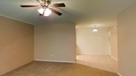This single-floor residence offers a straightforward and functional layout designed for comfortable everyday living. The main living area is finished with wall-to-wall carpeting in a warm neutral tone, providing a soft and inviting feel underfoot. The space is illuminated by a classic ceiling fan with an integrated light fixture, offering both ambient lighting and air circulation. The living area flows openly into an adjacent dining or secondary area, which is distinguished by a pendant light fixture hanging from the ceiling, creating a defined yet connected zone within the open floor plan. The walls throughout are finished in a soft beige tone, contributing to a neutral and versatile palette that complements a variety of décor styles. White baseboards provide a clean contrast along the perimeter of the rooms. The open passage between the two areas enhances the sense of space and allows for flexible use of the interior. Overall, the space presents a clean, move-in ready environment with consistent finishes throughout, making it well-suited for a range of furnishing arrangements and personal styles.